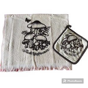 May include: Off-white kitchen towel and pot holder set featuring a mushroom design. The towel has a fringed edge and the words "Fresh Mushrooms" printed on it. The pot holder has a brown polka dot border and the words "Meadow Mushrooms".