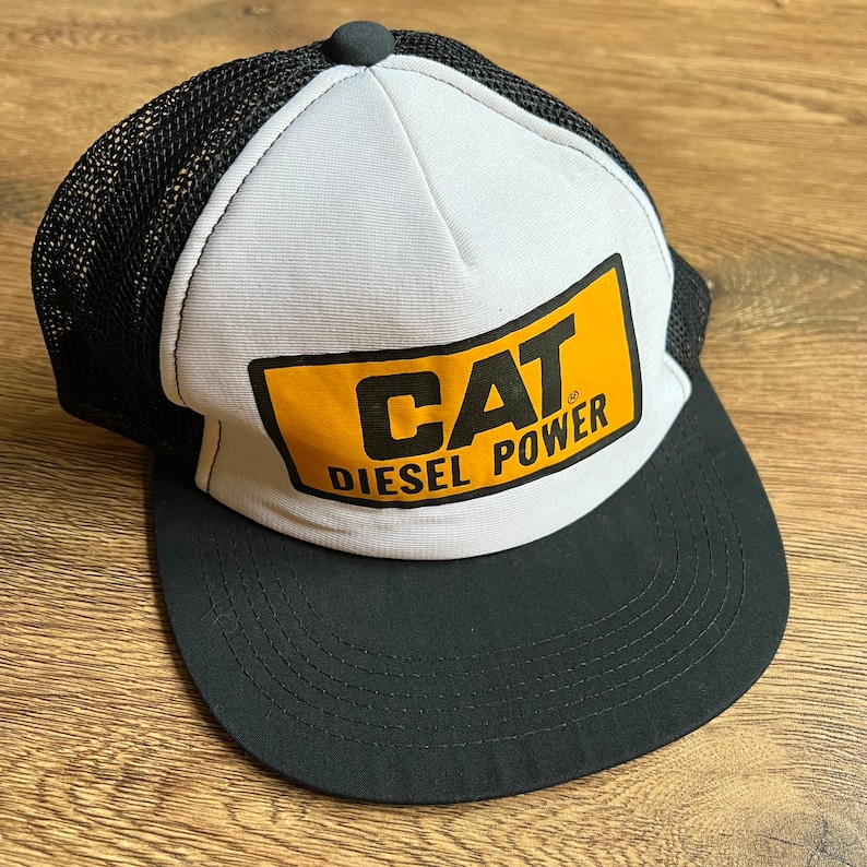 May include: A black and white trucker hat with a curved brim. The front panel is white with a yellow rectangle featuring the text "CAT DIESEL POWER" in black. The mesh back is black, and the hat has a black button on top.