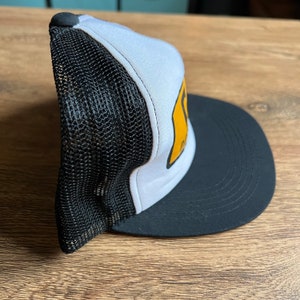 May include: A trucker hat with a black mesh back, a white front panel, and a black brim. The front panel features a yellow graphic. The hat has a black button on top.