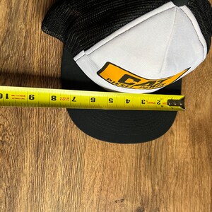 May include: Black and white trucker hat with a curved brim. The front panel is white with a yellow and black logo that reads "CAT DIESEL POWER." The brim measures approximately 9 inches.