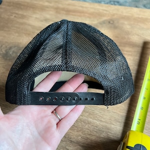May include: Black trucker hat with a mesh back and adjustable snap closure. The hat has a curved brim and a black front panel. The hat is designed to be worn with the brim facing forward.