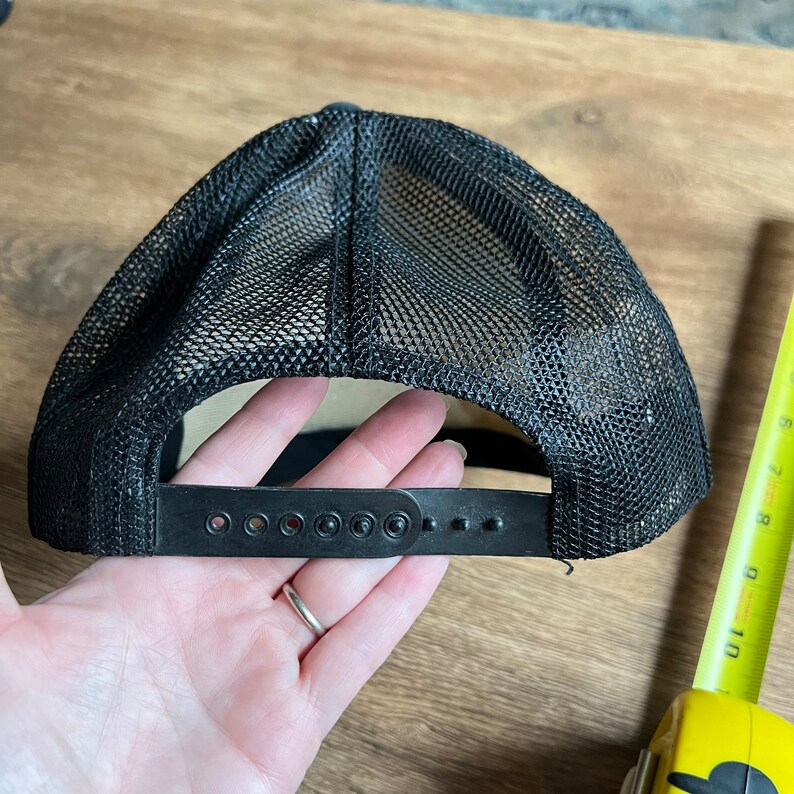 May include: Black trucker hat with a mesh back and adjustable snap closure. The hat has a curved brim and a black front panel. The hat is designed to be worn with the brim facing forward.