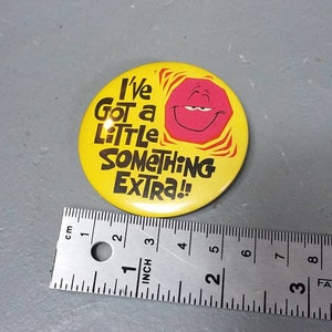 May include: A yellow button with the text "I've Got a Little Something Extra!!" in black. It features a smiling pink geometric shape. A ruler is placed next to the button, measuring in inches and centimeters.