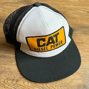 May include: A black and white trucker hat with a curved brim. The front panel is white with a yellow rectangle featuring the text "CAT DIESEL POWER" in black. The mesh back is black, and the hat has a black button on top.