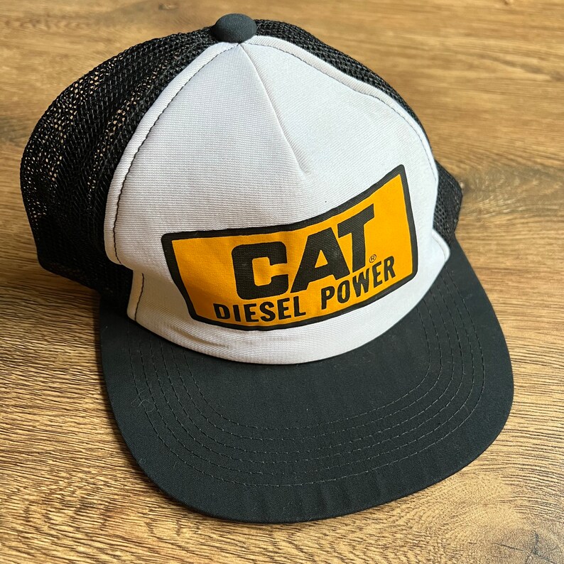 May include: A black and white trucker hat with a curved brim. The front panel is white with a yellow rectangle featuring the text "CAT DIESEL POWER" in black. The mesh back is black, and the hat has a black button on top.