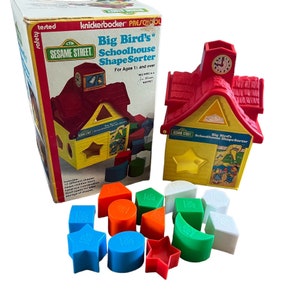 May include: A vintage Sesame Street Big Bird's Schoolhouse Shape Sorter toy. The yellow schoolhouse has a red roof and a clock tower. Includes 12 colorful geometric shapes in blue, orange, green, red, and white. The box is also visible.