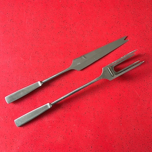 May include: A stainless steel carving set, including a long, slender knife with a pointed tip and a two-tined fork. The knife has a slightly curved blade. Both utensils have rectangular handles and are set against a red background.
