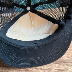 May include: Black trucker hat with a mesh back and a curved brim. The interior features a beige lining and a white button at the top. The hat's design includes a black sweatband and a structured front panel.