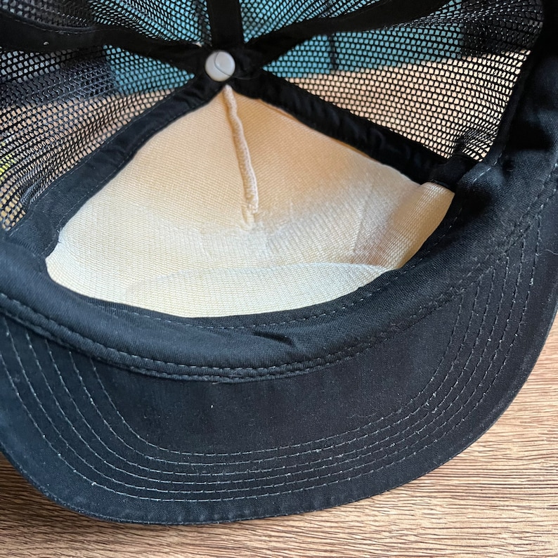 May include: Black trucker hat with a mesh back and a curved brim. The interior features a beige lining and a white button at the top. The hat's design includes a black sweatband and a structured front panel.