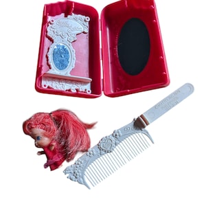 Puede incluir: Un juego de Sweet Secrets Salon vintage, con una caja roja con un espejo, una muñeca pequeña con pelo rojo y un peine azul claro. El espejo tiene una superficie brillante azul. El peine tiene un diseño floral decorativo.
