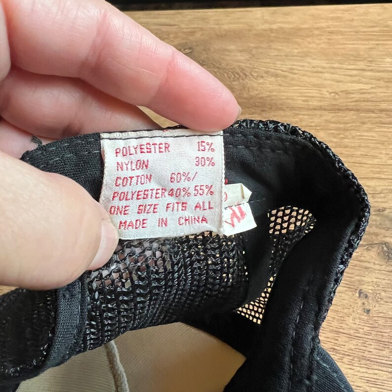 May include: A black mesh trucker hat with a tag showing the fabric composition: 15% polyester, 30% nylon, 60% cotton, and 40%/55% polyester. The tag also states "One Size Fits All" and "Made in China."