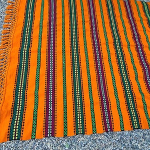 May include: An orange and striped textile with a fringed edge. The textile features vertical stripes in green, purple, and white, with a repeating pattern. The textile is likely a blanket or throw.