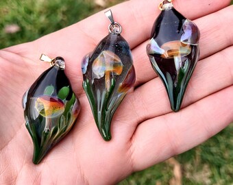 May include: Three glass mushroom pendants with gold chains. Each pendant features a detailed mushroom with a brown cap and white stem, surrounded by green leaves. The glass is clear and iridescent, creating a shimmering effect.