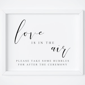 May include: A white framed sign with black script that reads "love is in the air" and "please take some bubbles for after the ceremony". The sign is a wedding or event decoration.