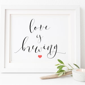 Puede incluir: Impresión enmarcada en blanco con caligrafía negra que dice "love is brewing" con un pequeño corazón rojo debajo del texto.