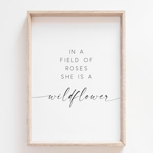 May include: A framed print with a white background and black text that reads "In a field of roses she is a wildflower".