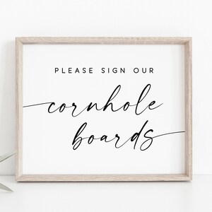 May include: A white sign in a light wood frame with black text that reads "Please sign our cornhole boards".