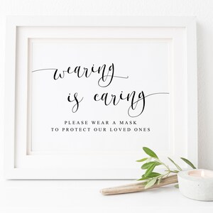 May include: White framed sign with black calligraphy that reads "wearing is caring" and the text "Please wear a mask to protect our loved ones".