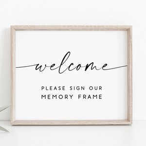 Könnte beinhalten: Ein weißer gerahmter Schild mit schwarzem Text, der "welcome" in Kursivschrift und "Please sign our memory frame" in einer serifenlosen Schriftart lautet.