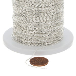 Puede incluir: Un carrete de cadena plateada, con una pequeña sección de la cadena dispuesta junto a un céntimo de euro para la escala. La cadena está hecha de pequeños eslabones rectangulares.