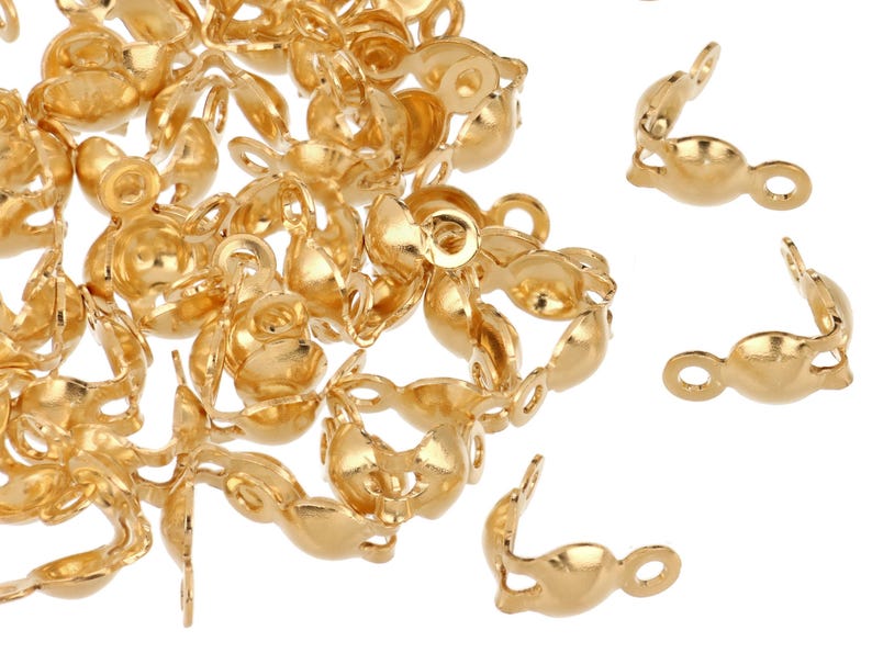 May include: A collection of small, gold-colored jewelry findings, including crimp end caps and connectors. The findings are made of a shiny, metallic material and are scattered across a white surface. The end caps have a curved shape with a loop.
