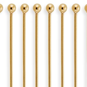 May include: Ten gold-colored metal pins with round heads. The pins are arranged in a row.