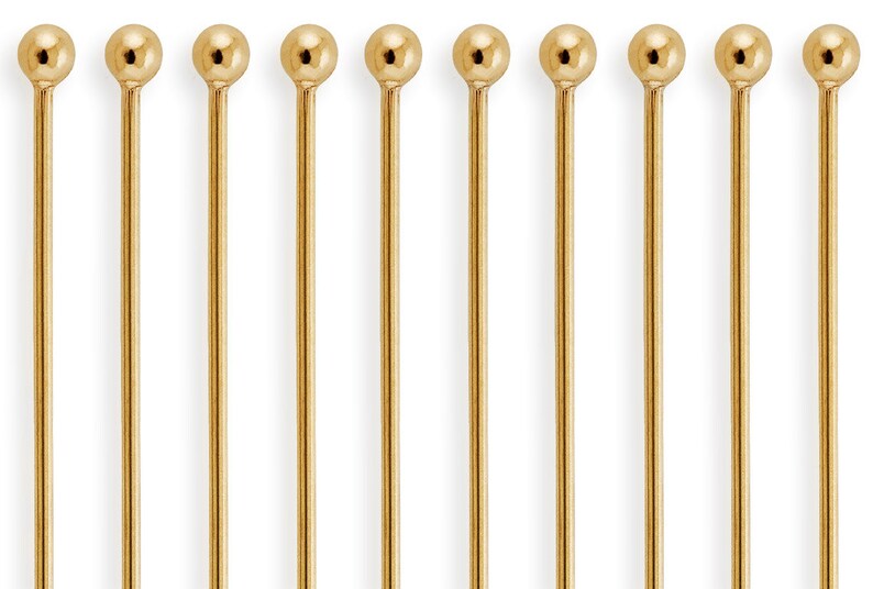 May include: Ten gold-colored metal pins with round heads. The pins are arranged in a row.