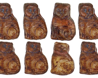 Może przedstawiać: Osiem małych, brązowych ceramicznych figurek kotów. Każdy kot siedzi z ogonem owiniętym wokół nóg. Kotki mają szkliwioną powierzchnię, a niektóre mają niebieskie oczy.