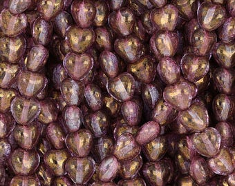May include: A close-up of a pile of heart-shaped glass beads. The beads are a translucent purple color with gold flecks.