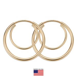Op de afbeelding: Een paar goudkleurige oorringen. De oorbellen zijn cirkelvormig, met een kleinere ring genest in een grotere. De afbeelding bevat een kleine Amerikaanse vlag en de tekst "MADE IN USA".