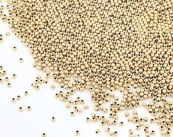 May include: A large pile of gold-colored round beads. The beads are scattered on a white surface.
