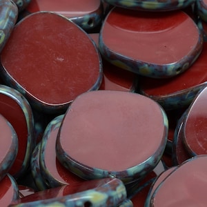 May include: Close-up of a collection of maroon-colored, irregularly shaped beads. Each bead has a glossy, smooth surface with a teal and yellow-green rim. The beads are slightly rounded and have a hole for stringing.