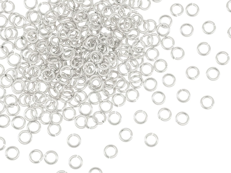 May include: A large pile of silver jump rings, with a few scattered around the pile. The jump rings are small and circular, with a gap in the circle.