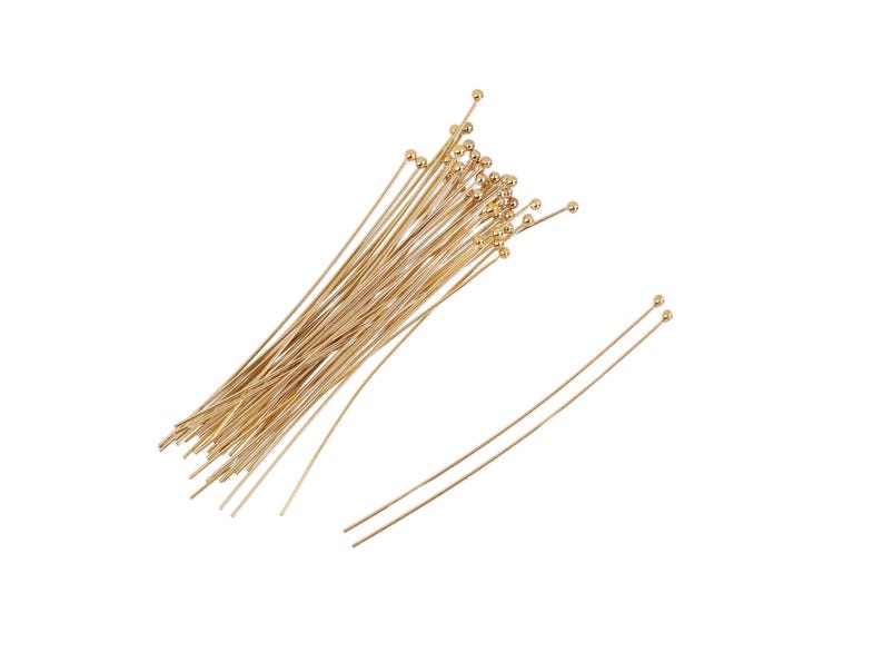 May include: Gold headpins, a type of jewelry finding, are shown in two orientations.  A bundle of approximately 30 gold headpins is lying on a white background.  A few individual headpins are lying separately. The headpins have small, round gold heads and slender, straight, gold shafts. These are ideal for jewelry making and crafting.