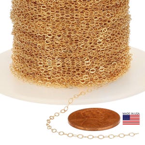 May include: A spool of gold-colored chain with oval links. A small section of the chain is unraveled, next to a penny for size comparison. The chain is on a white surface. The American flag and the text "MADE IN USA" are in the lower right corner.