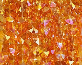 May include: Close-up of a collection of orange heart-shaped beads. The beads are faceted, catching the light and creating a sparkling effect. The beads are a warm, vibrant orange colour with hints of pink and yellow.