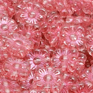 May include: A close-up shot of numerous translucent pink flower-shaped beads. Each bead has a central raised circle and petal-like edges. The beads are clustered together, creating a textured, floral pattern. The beads are made of a shiny, transparent material.
