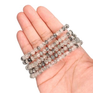 Puede incluir: Varias hebras de cuentas de piedras preciosas claras y grises, probablemente cuarzo, se sostienen en una mano. Las peque&ntilde;as cuentas redondas tienen un aspecto moteado. Las cuentas est&aacute;n ensartadas, formando m&uacute;ltiples hebras.