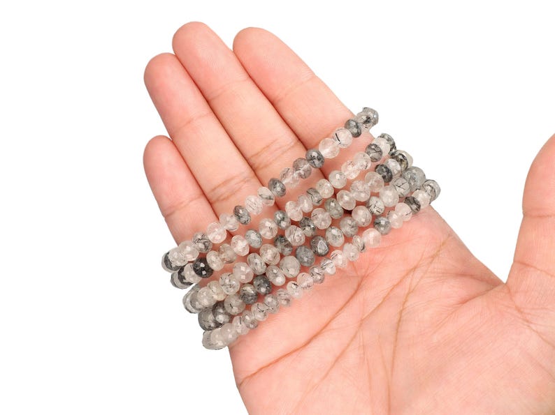 Puede incluir: Varias hebras de cuentas de piedras preciosas claras y grises, probablemente cuarzo, se sostienen en una mano. Las peque&ntilde;as cuentas redondas tienen un aspecto moteado. Las cuentas est&aacute;n ensartadas, formando m&uacute;ltiples hebras.