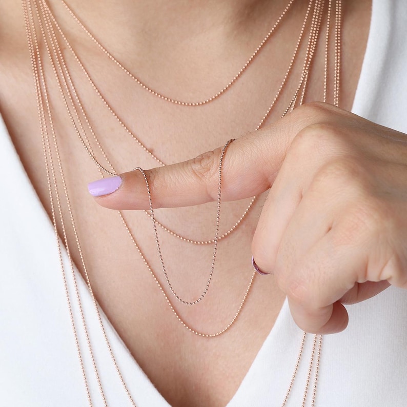 May include: Several delicate, rose gold ball chain necklaces of varying lengths are displayed. One chain is held by a finger with a light purple nail polish. The necklaces are layered, creating a cascading effect against a white shirt.