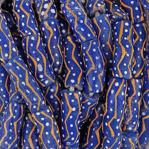 Puede incluir: Primer plano de una colección de cuentas de vidrio azul con patrones blancos y naranjas. Las cuentas son cilíndricas y tienen una superficie texturizada.