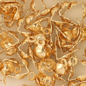 May include: A collection of small, gold-colored jewelry findings, including crimp end caps and triangle-shaped components. The findings have a shiny, metallic finish and are scattered across a light-colored surface. These components are used in jewelry making.