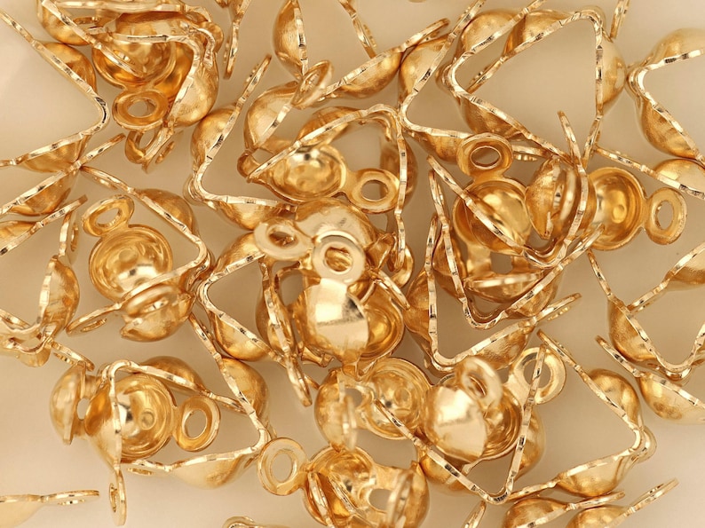 May include: A collection of small, gold-colored jewelry findings, including crimp end caps and triangle-shaped components. The findings have a shiny, metallic finish and are scattered across a light-colored surface. These components are used in jewelry making.