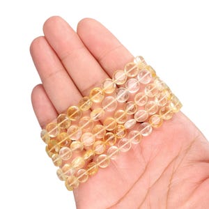 May include: A stack of four bracelets made of translucent, golden-yellow citrine beads. The beads are round and strung together on a clear elastic cord. The bracelets are displayed on a light skin tone background.