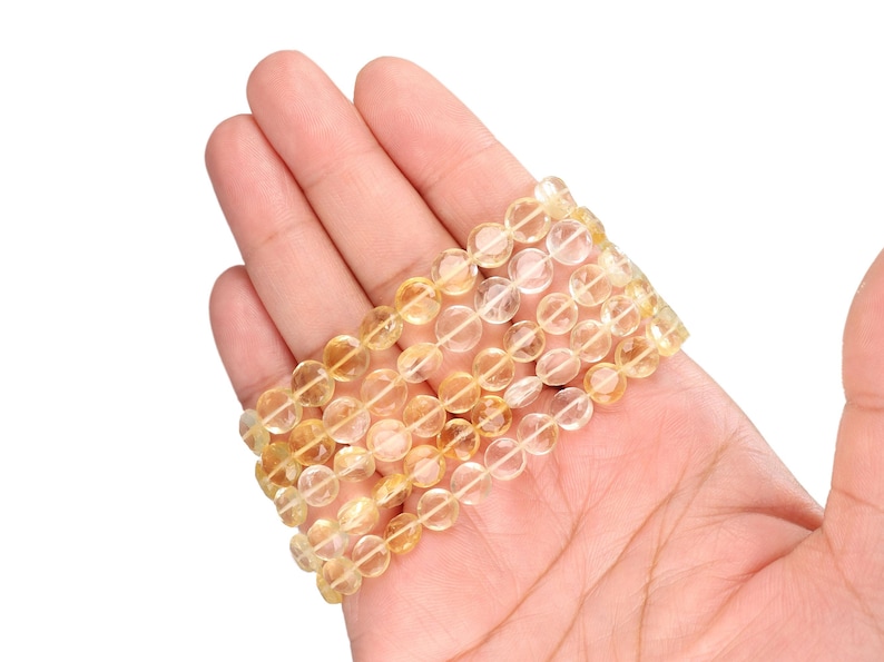 May include: A stack of four bracelets made of translucent, golden-yellow citrine beads. The beads are round and strung together on a clear elastic cord. The bracelets are displayed on a light skin tone background.