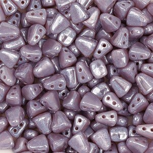 May include: A close-up of a pile of small, iridescent, light purple triangle-shaped beads. The beads have two holes each.