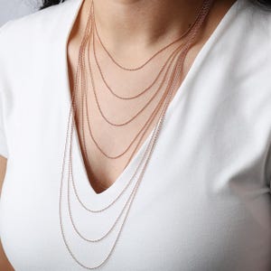 May include: A layered necklace with multiple strands of delicate, rose gold-colored chains. The chains are composed of small, round beads and cascade down the chest, creating a stylish and elegant look. The necklace is worn over a white v-neck shirt.