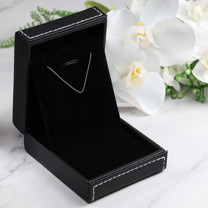 May include: Open black jewelry box with a gold chain necklace displayed inside. The box is rectangular with white stitching around the edges. White flowers and greenery are in the background.