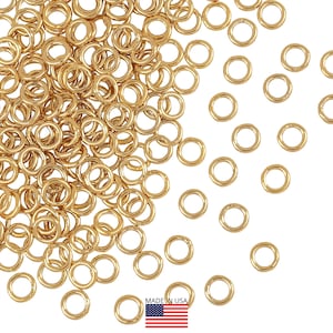 May include: A collection of small, gold-coloured jump rings scattered on a white surface. The rings are circular and appear to be made of metal. The image includes a small "Made in USA" label with an American flag.