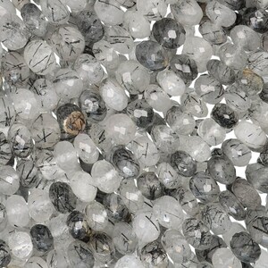 Puede incluir: Un primer plano de una colecci&oacute;n de cuentas de piedras preciosas claras y facetadas con inclusiones negras. Las cuentas de forma ovalada var&iacute;an en tonos de gris y claro, creando una apariencia texturizada. Ideal para la creaci&oacute;n de joyas.